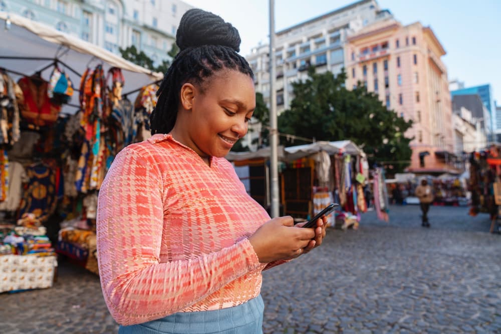 Woman in a colorful market square smiling while using her smartphone, surrounded by local street stalls.