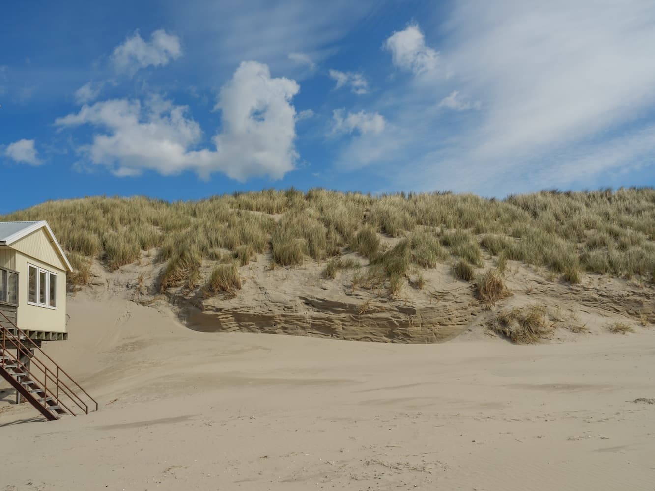 A raised beach hut on wooden stilts before towering sand dunes, its pale exterior contrasting with driftwood and ocean-washed sands under a blue sky.