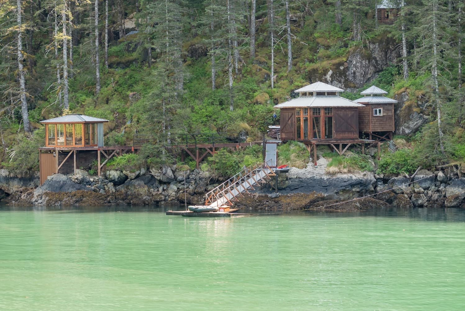 Two eco-pods and a glass igloo nestled on a rocky lakeshore amid dense native forest, with a small jetty leading into emerald-green waters.