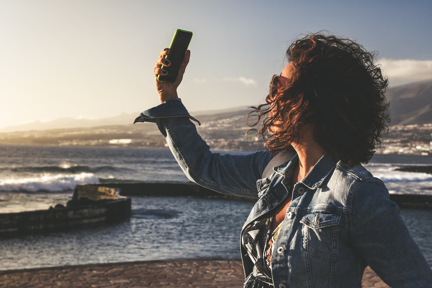 A woman stands on a black-sand beach at sunset, holding her phone aloft to capture the ocean and distant hills behind her.