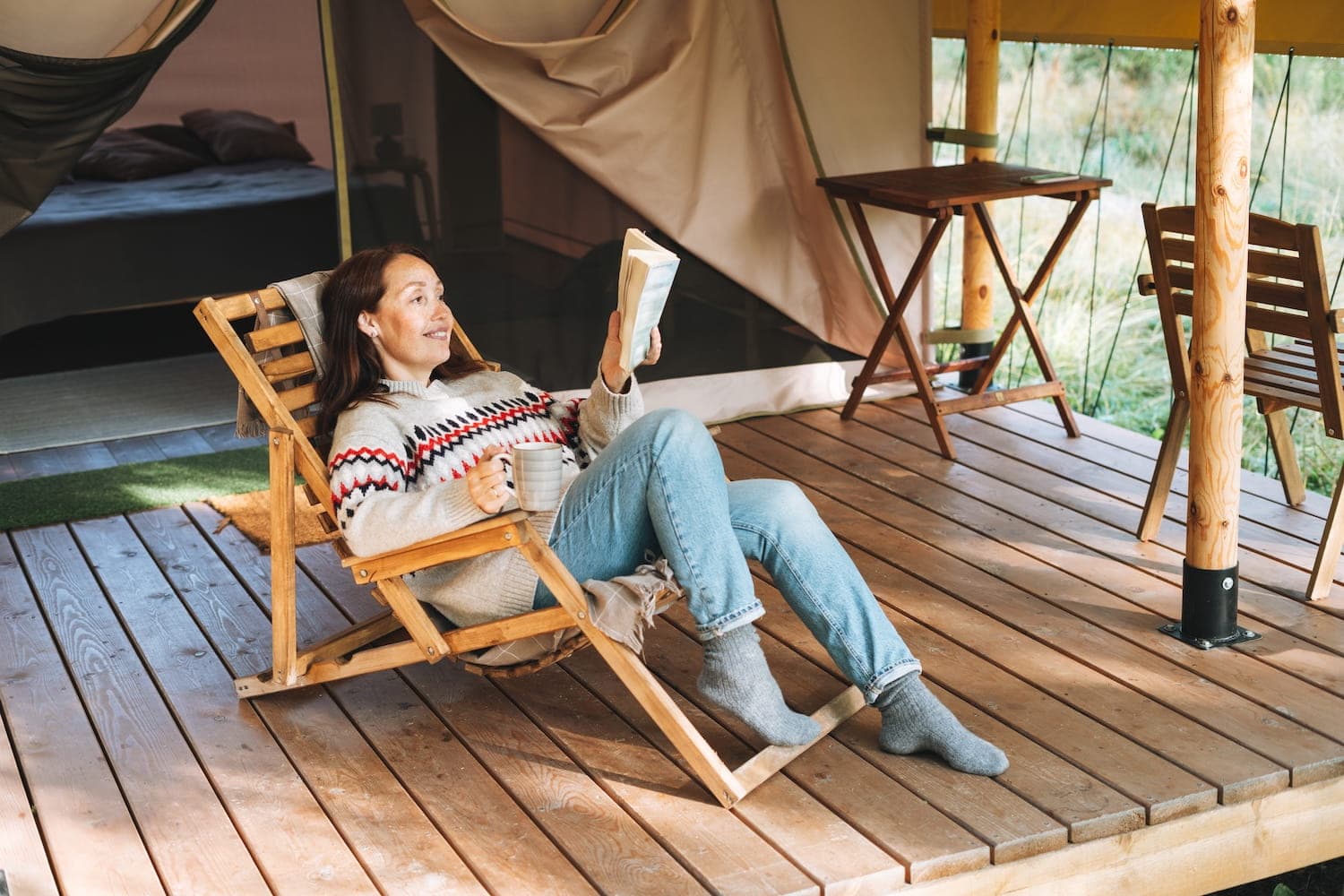 A woman relaxing in a folding lounge chair on the deck of a safari-style tent, reading a book as sunlight filters through surrounding trees.