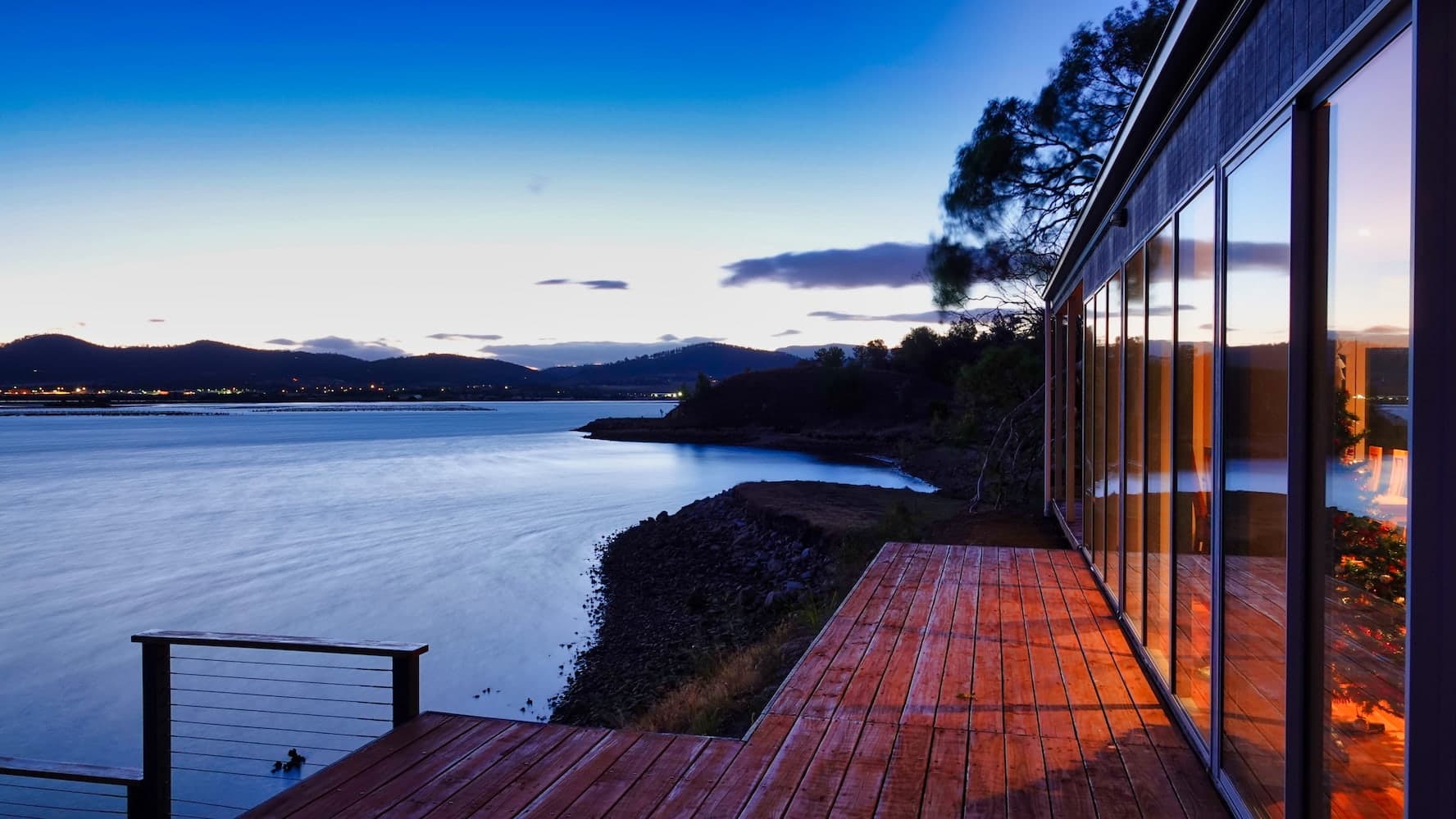 A modern deck with full-height glass windows overlooking a deep blue bay at dusk, framed by rocky shore and silhouetted hills.