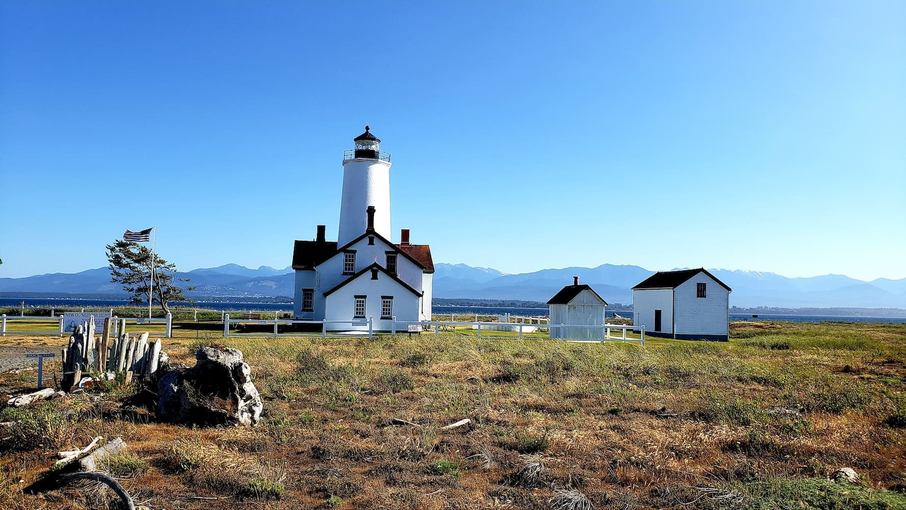 A white lighthouse and keeper’s cottage standing on windswept grassland beside a rugged shoreline with distant mountain silhouettes.