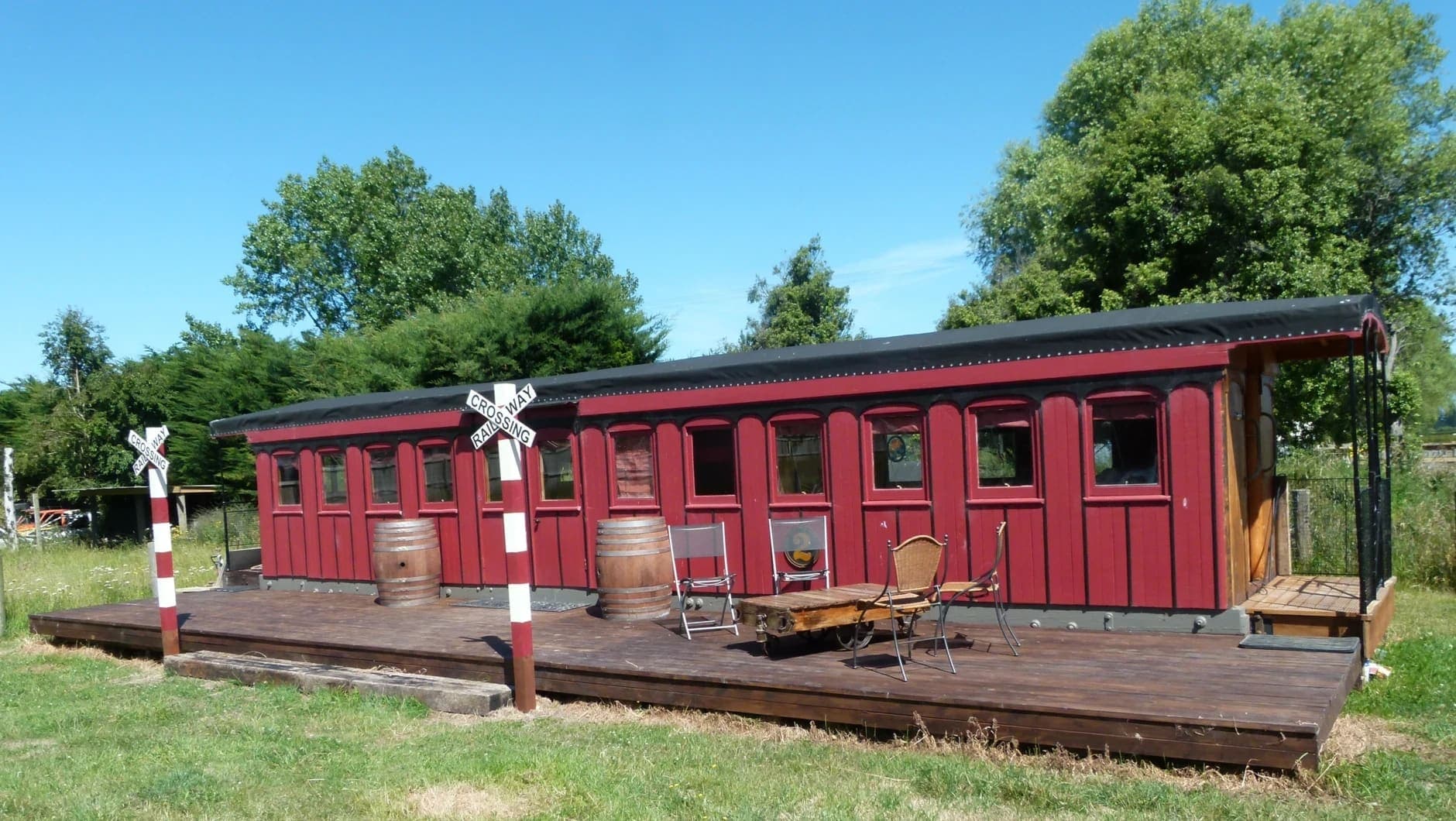 A refurbished red railway carriage converted into a hotel room, sitting on rails in a grassy field with a veranda at one end.