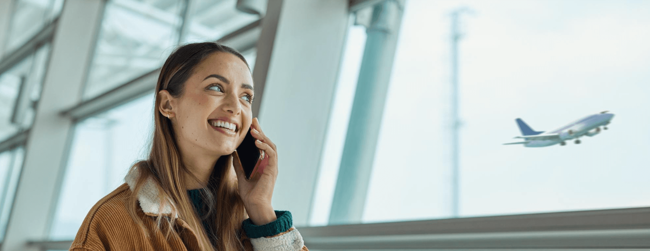 Woman smiling while talking on the phone at an airport, with a plane taking off in the background.