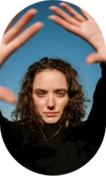 Person with curly hair and a nose ring, wearing a black top, raises hands against a clear blue sky backdrop.