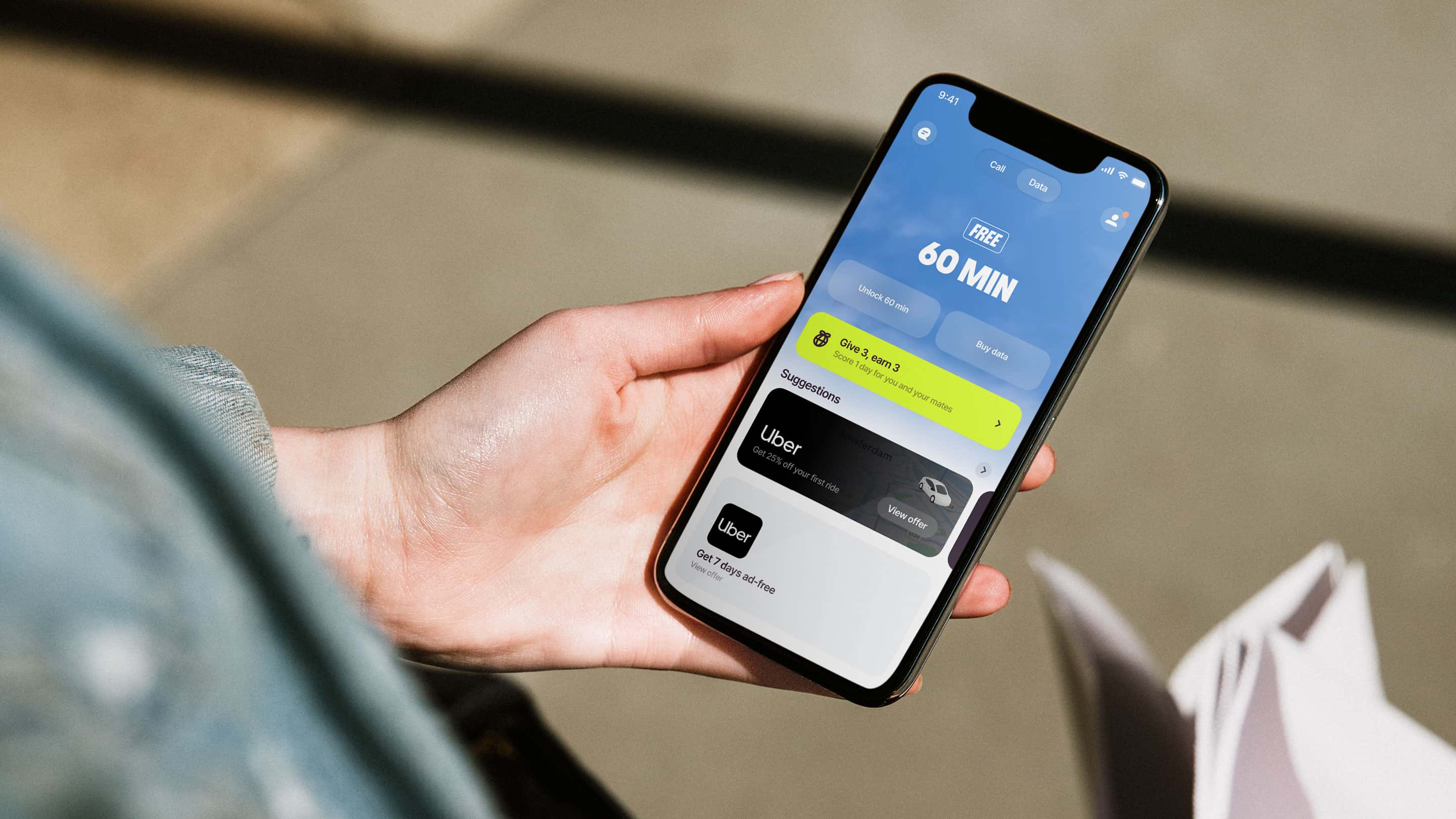 A hand holding a smartphone displaying the Firsty app interface, showing a screen with "FREE 60 MIN" of data, options to unlock more time or buy data, a referral offer, and Uber ride suggestions; the phone rests on a desk with notebooks and a wooden object nearby.