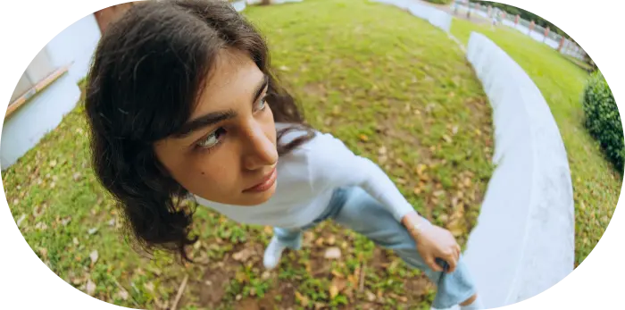 Person with long hair in a white shirt and jeans, captured with a fisheye lens, standing on grass near a curved path, looking to the side.