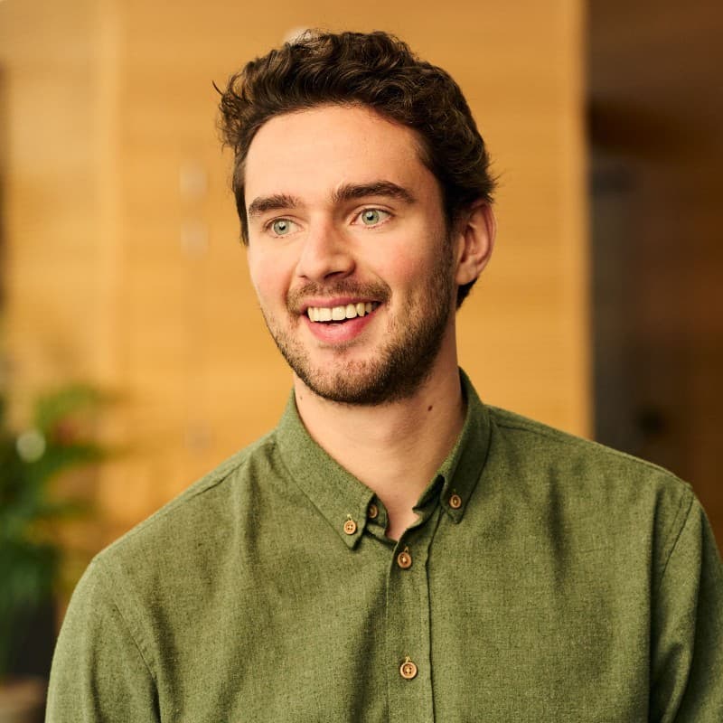 Smiling man with short curly hair and a beard, wearing a green button-up shirt, stands indoors with a blurred background.