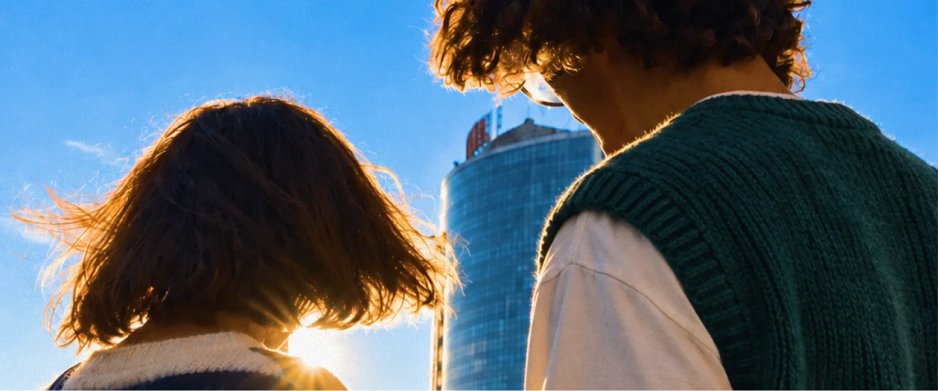 Two colleagues from behind on a rooftop, the low sun behind a glass tower between them