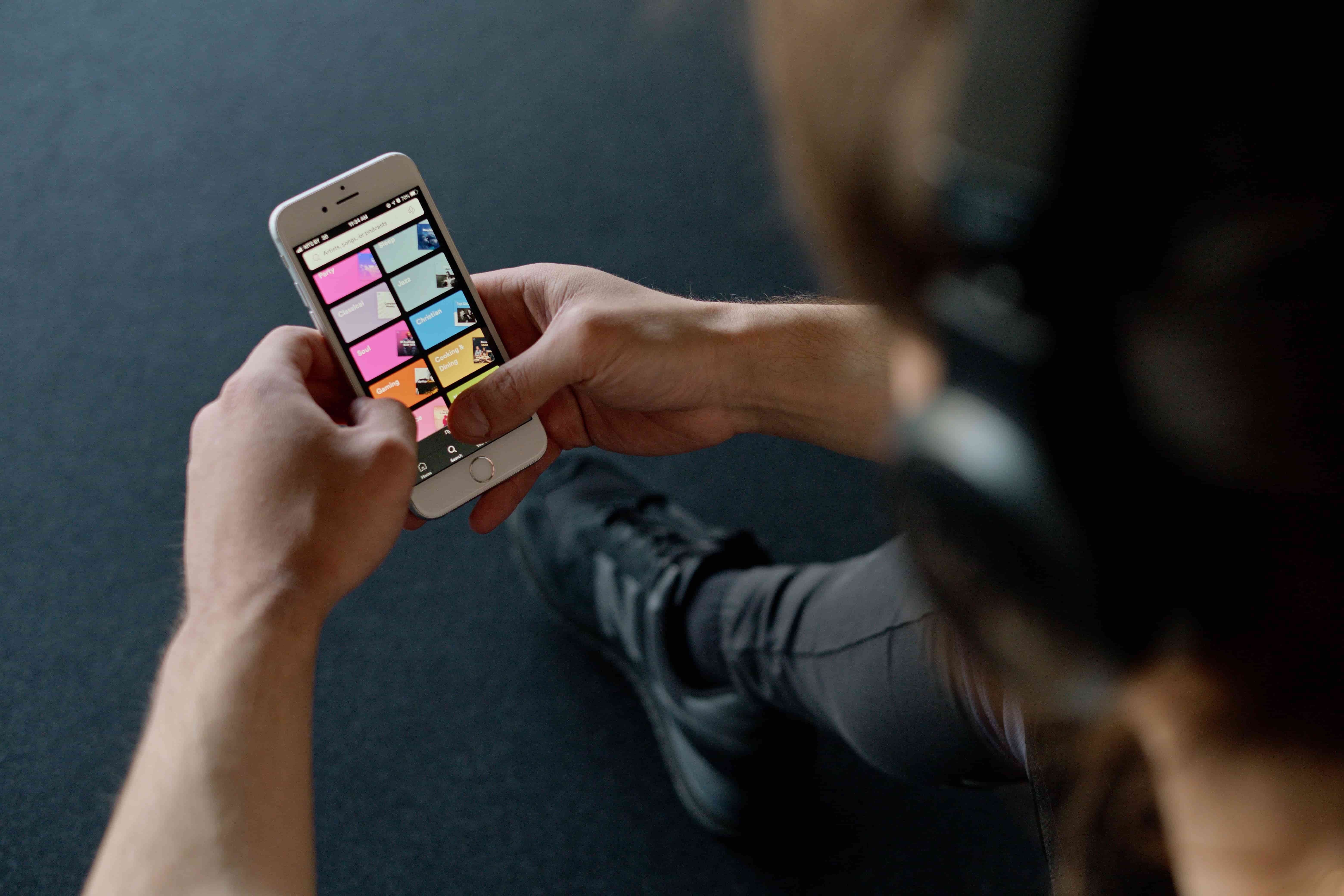 Person wearing headphones using a smartphone with a Spotify app interface while sitting on a dark carpeted floor.